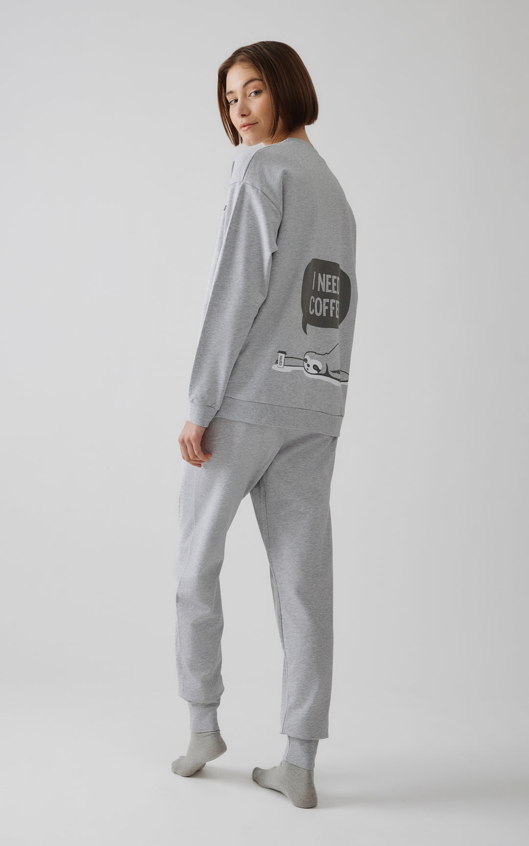 Πίσω όψη γυναίκας με γκρι πιτζάμα με στάμπα cartoon τεμπέλη και φράση "I need coffee" στο πίσω μέρος.