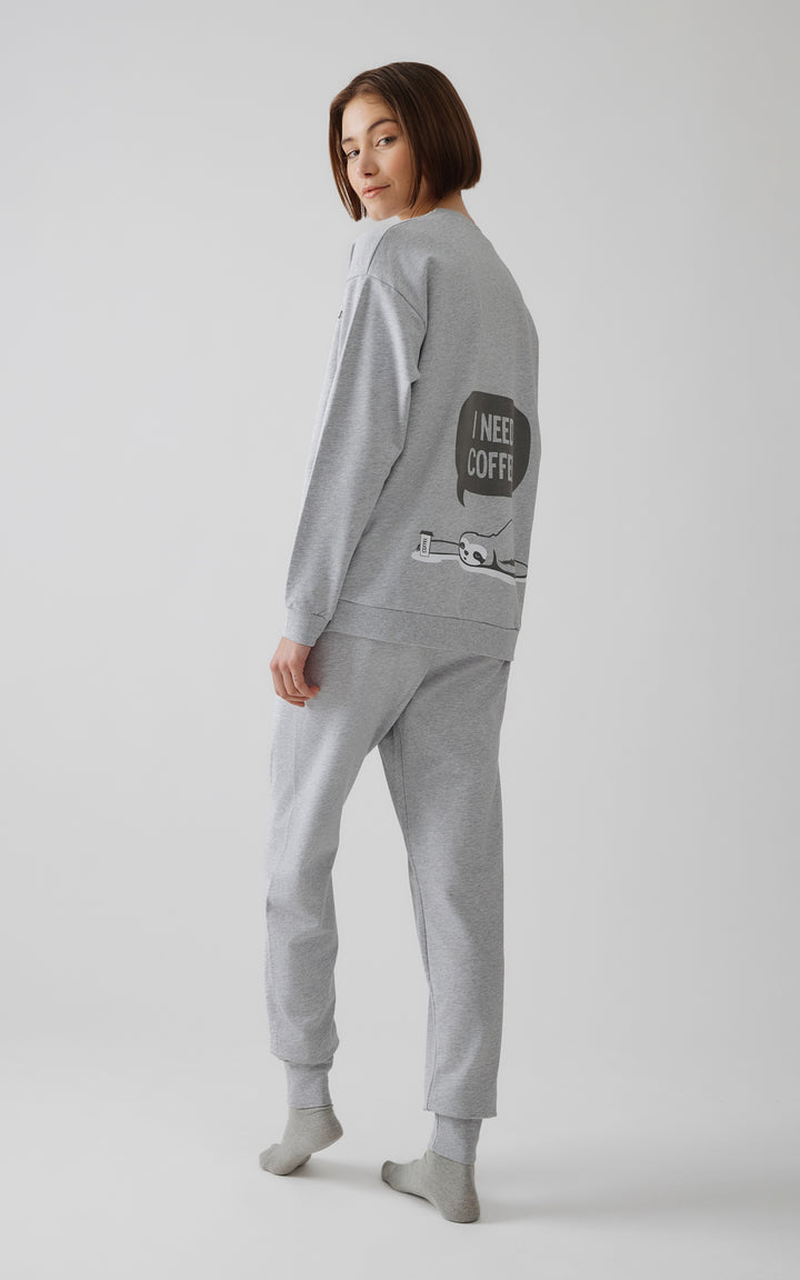 Πίσω όψη γυναίκας με γκρι πιτζάμα με στάμπα cartoon τεμπέλη και φράση "I need coffee" στο πίσω μέρος.