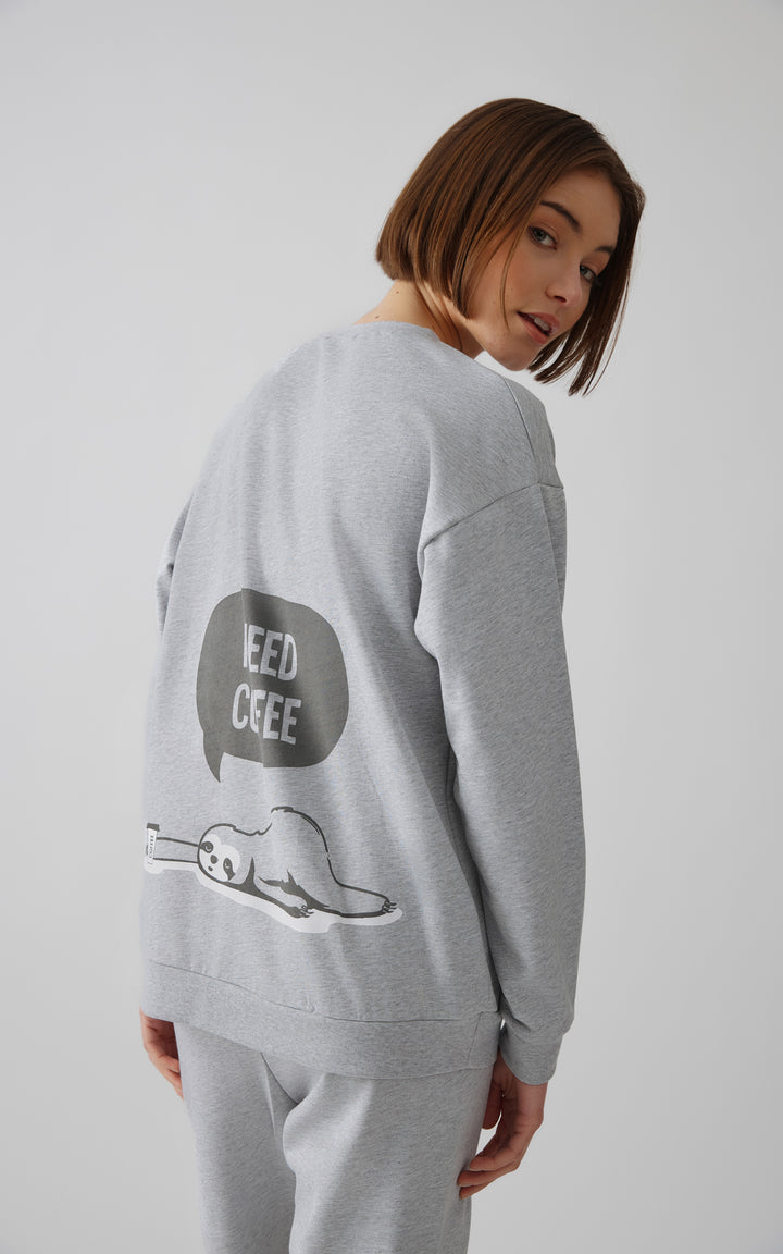 Γυναίκα με καρέ καστανά μαλλιά φορά γκρι πιτζάμα και γυρνά ελαφρώς το κεφάλι, δείχνοντας τη στάμπα με τεμπέλη που γράφει "I need coffee".