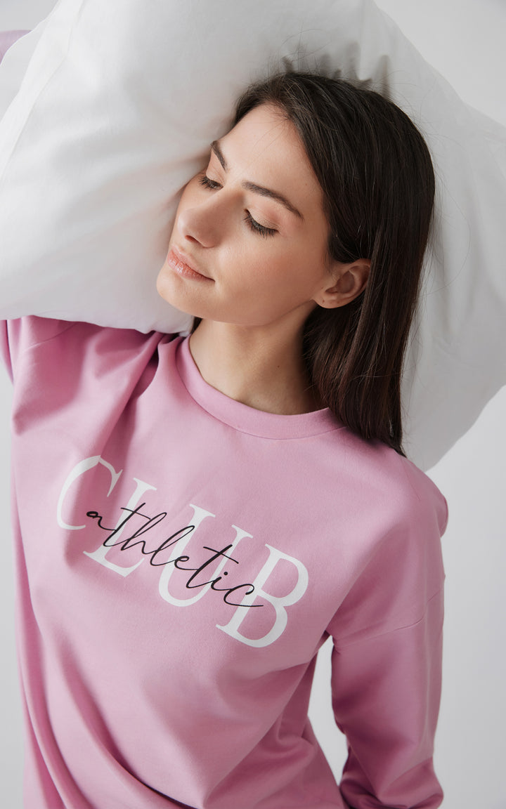 Γυναικείο μοντέλο με μακριά μαλλιά ξαπλώνει σε λευκό μαξιλάρι με κλειστά μάτια, φορώντας ροζ homewear μπλούζα με τύπωμα "athletic CLUB", αποπνέοντας αίσθηση άνεσης και χαλάρωσης.