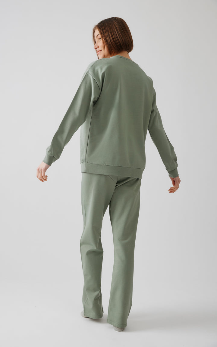 Γυναικείο μοντέλο ποζάρει χαμογελώντας διακριτικά με γυρισμένη πλάτη, φορώντας χακί homewear σετ, σε καθαρό και φωτεινό περιβάλλον με γκρι background.