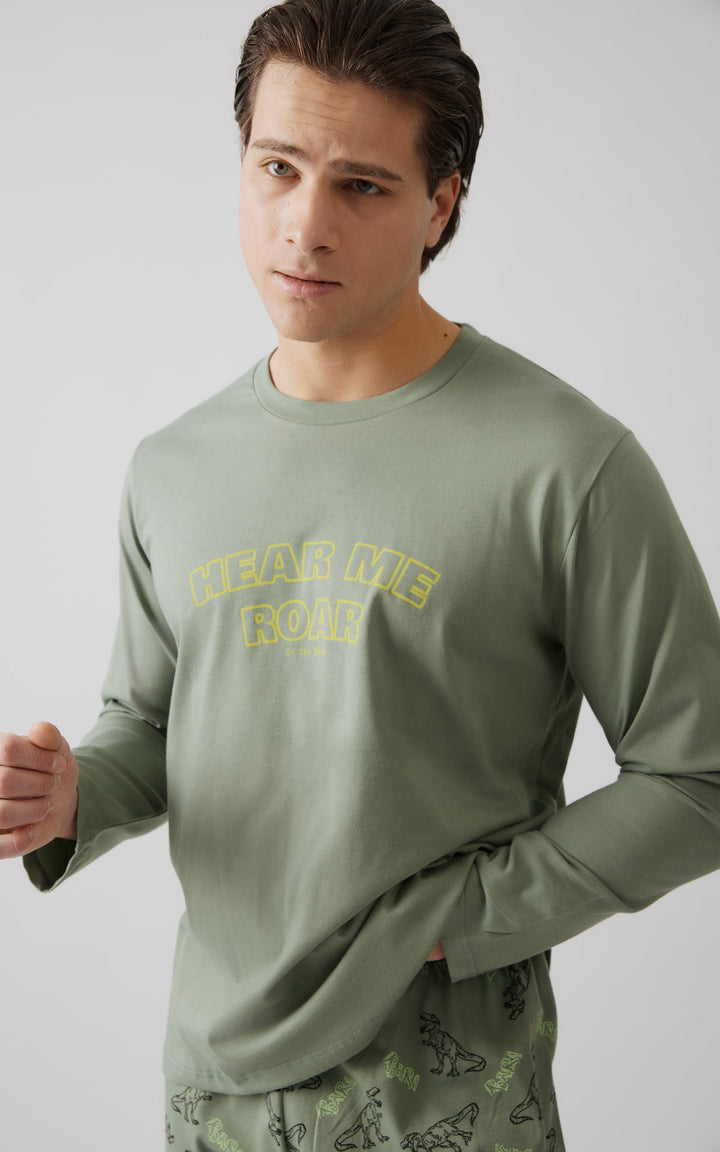 Άνδρας σε κοντινή λήψη ποζάρει με ήπια έκφραση, φορώντας χακί μακρυμάνικη μπλούζα πιτζάμας με κίτρινη στάμπα “Hear Me Roar” και ασορτί παντελόνι με μοτίβα δεινοσαύρων.