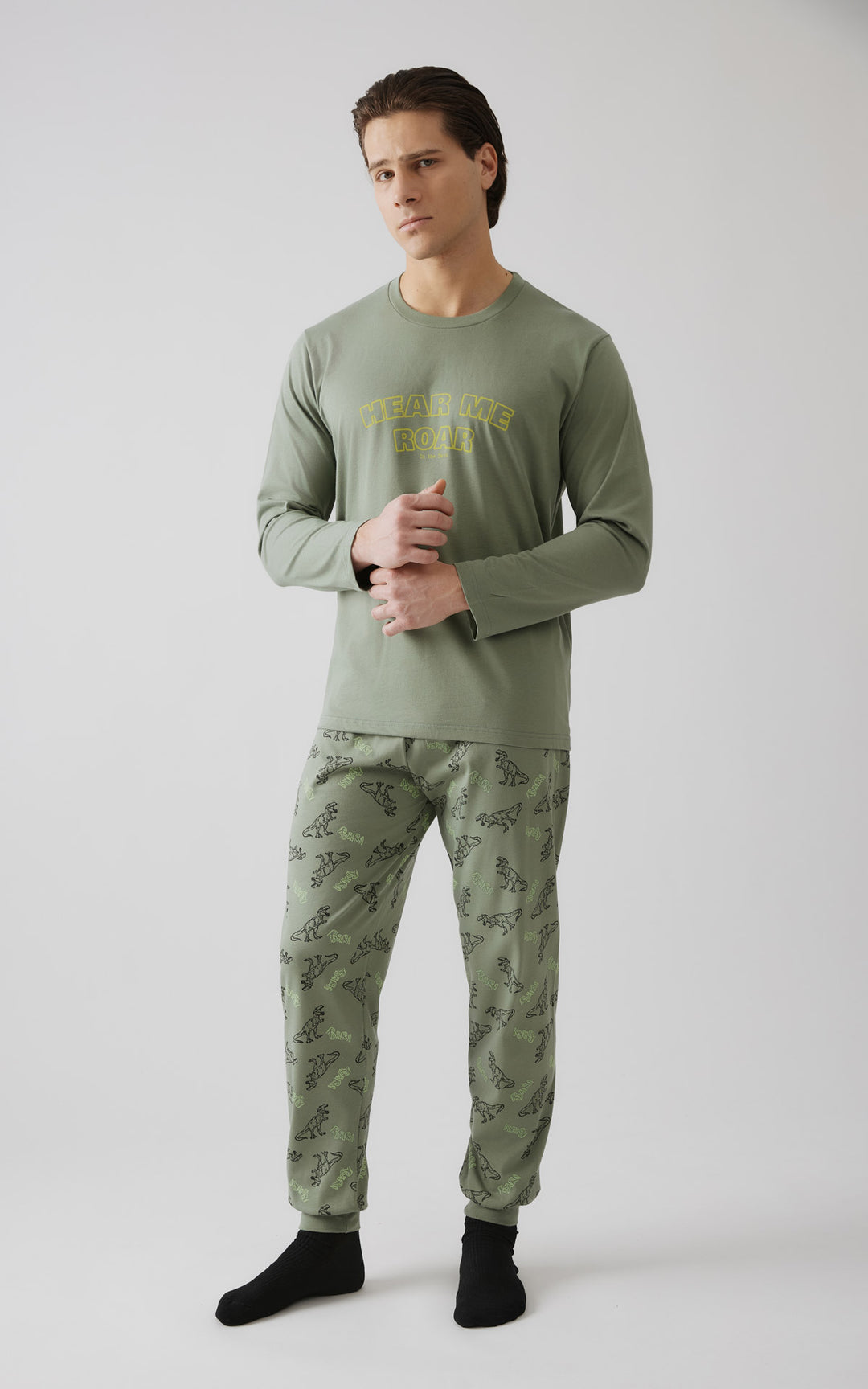 Ανδρικό μοντέλο  με σταυρωμένα χέρια φοράει χακί σετ πιτζάμας με μακρυμάνική μπλούζα και παντελόνι με σχέδιο δεινοσαύρων.