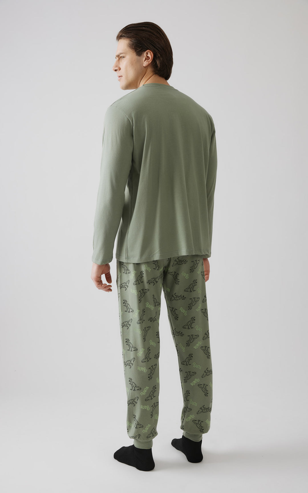 Άνδρας με πλάγια στάση γυρισμένος προς τα πίσω, φοράει σετ με χακί πιτζάμες με σχέδιο με δεινόσαυρους και μαύρες κάλτσες. 