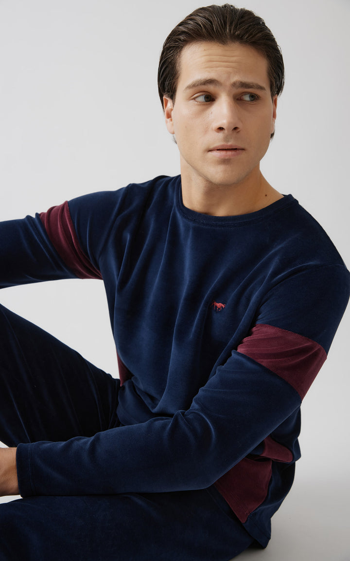 Άντρας φοράει βελουτέ μπλε πιτζάμα με μπορντό λεπτομέρειες στα μανίκια και το πλάι, καθισμένος στο πάτωμα σε χαλαρή στάση.