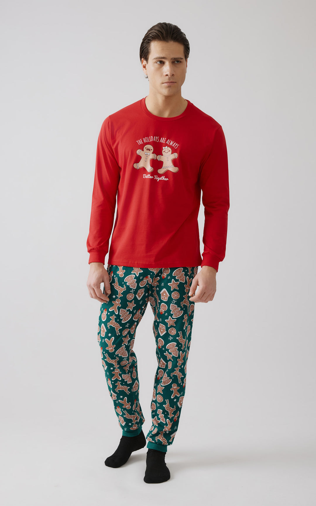 Ολόσωμη εικόνα ανδρικού μοντέλου που φοράει Χριστουγεννιάτικη πιτζάμα με κόκκινη μπλούζα και παντελόνι γεμάτο μοτίβα από μπισκότα gingerbread.