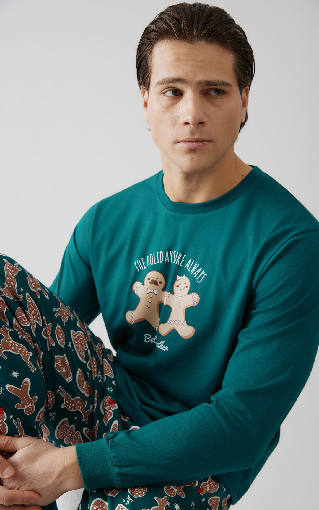 Άνδρας καθισμένος σε στούντιο φοράει πράσινη πιτζάμα με χριστουγεννιάτικο σχέδιο. Η μπλούζα έχει στάμπα με δύο μπισκότα gingerbread και τη φράση "The holidays are always better together".