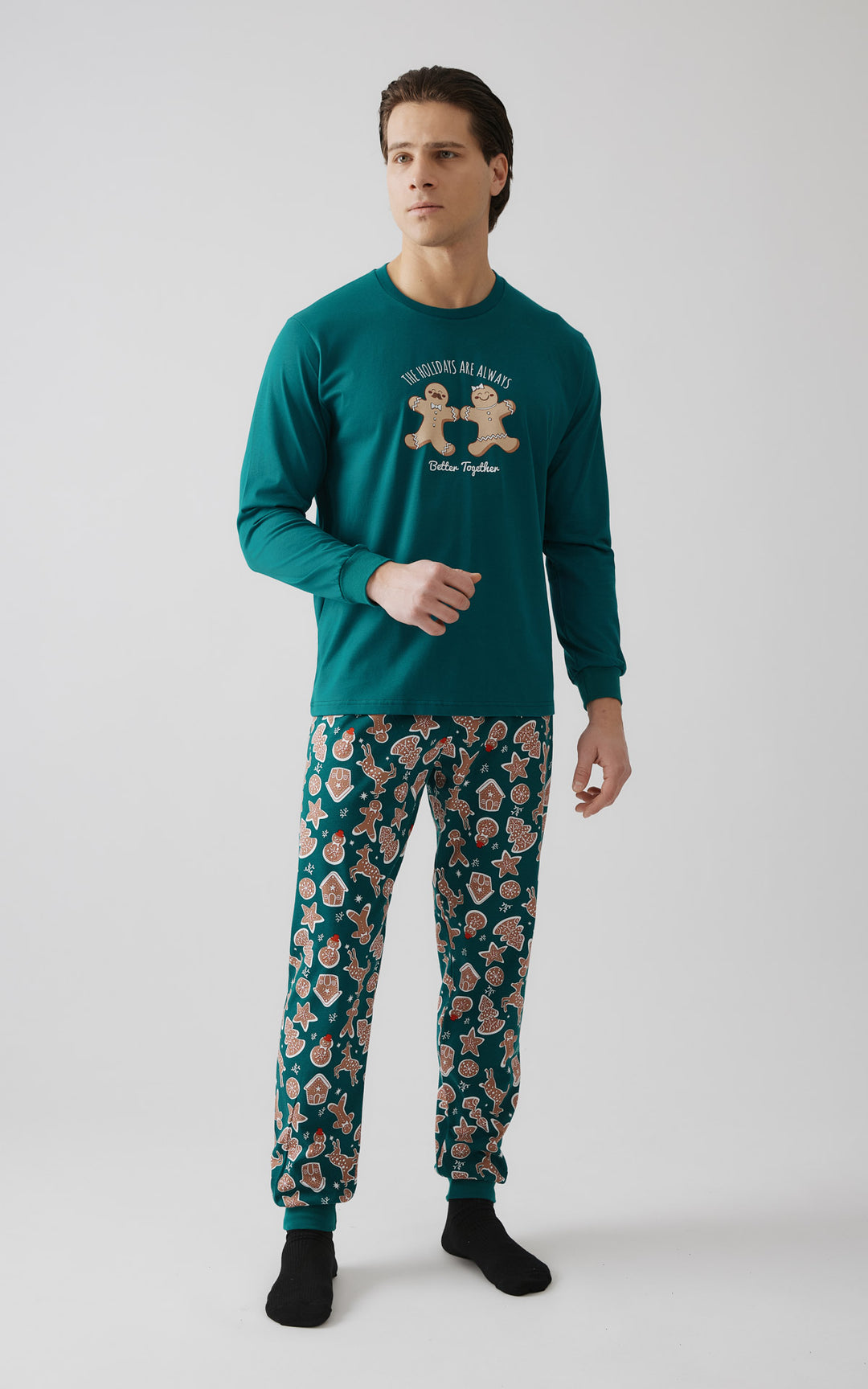 Άνδρας όρθιος  κοιτάζοντας στο πλάι και φορώντας πράσινη χριστουγεννιάτικη πιτζάμα με στάμπα μπισκότα στη μπλούζα και μοτίβο gingerbread σε όλο το παντελόνι.