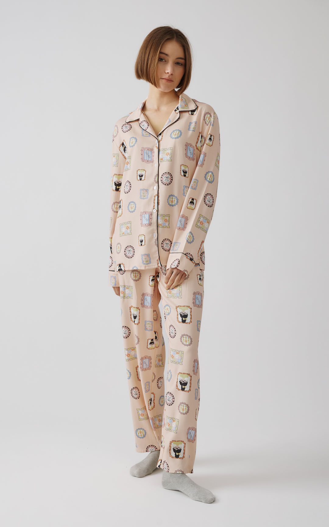 Γυναικείο μοντέλο στέκεται χαλαρά με απαλή ροζ πιτζάμα γεμάτη πολύχρωμα σχέδια  με κάδρα και μαύρες λεπτομέρειες στο γιακά και τα μανίκια, κοιτάζοντας την κάμερα με απαλή έκφραση σε ουδέτερο περιβάλλον.