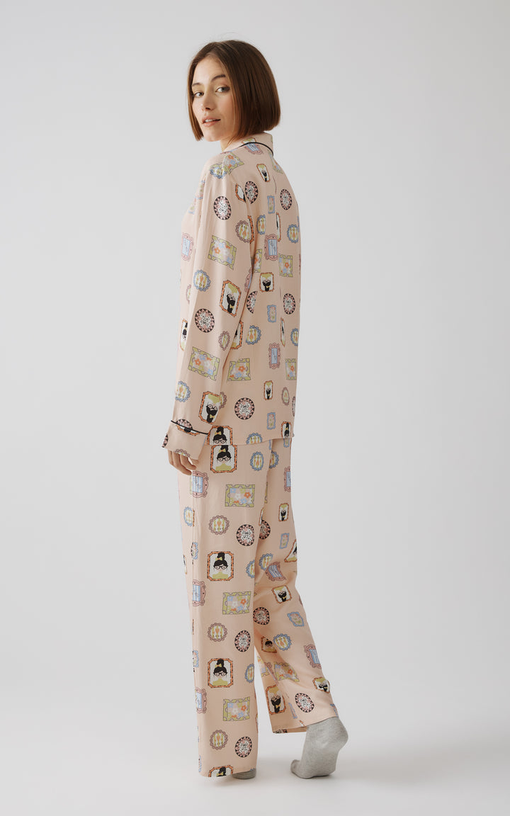 Γυναίκα ποζάρει γυρισμένη ελαφρώς προς τα πίσω φορώντας ροζ πιτζάμα με παιχνιδιάρικο μοτίβο από πολύχρωμα καρέ και φιγούρες, χαμογελώντας διακριτικά σε φωτεινό στούντιο με λευκό φόντο.