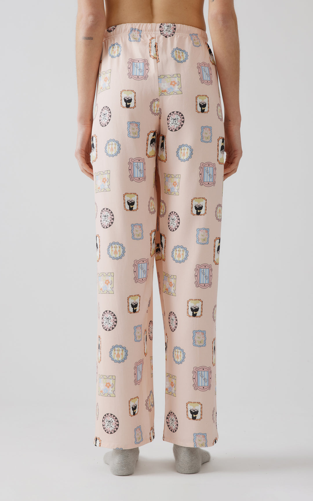 Η πίσω εικόνα γυναικείου μοντέλου από τη μέση και κάτω που φοράει μαύρη μακρυμάνικη μπλούζα και παντελόνι πιτζάμα με πολύχρωμα σχέδια και γκρι κάλτσες. 