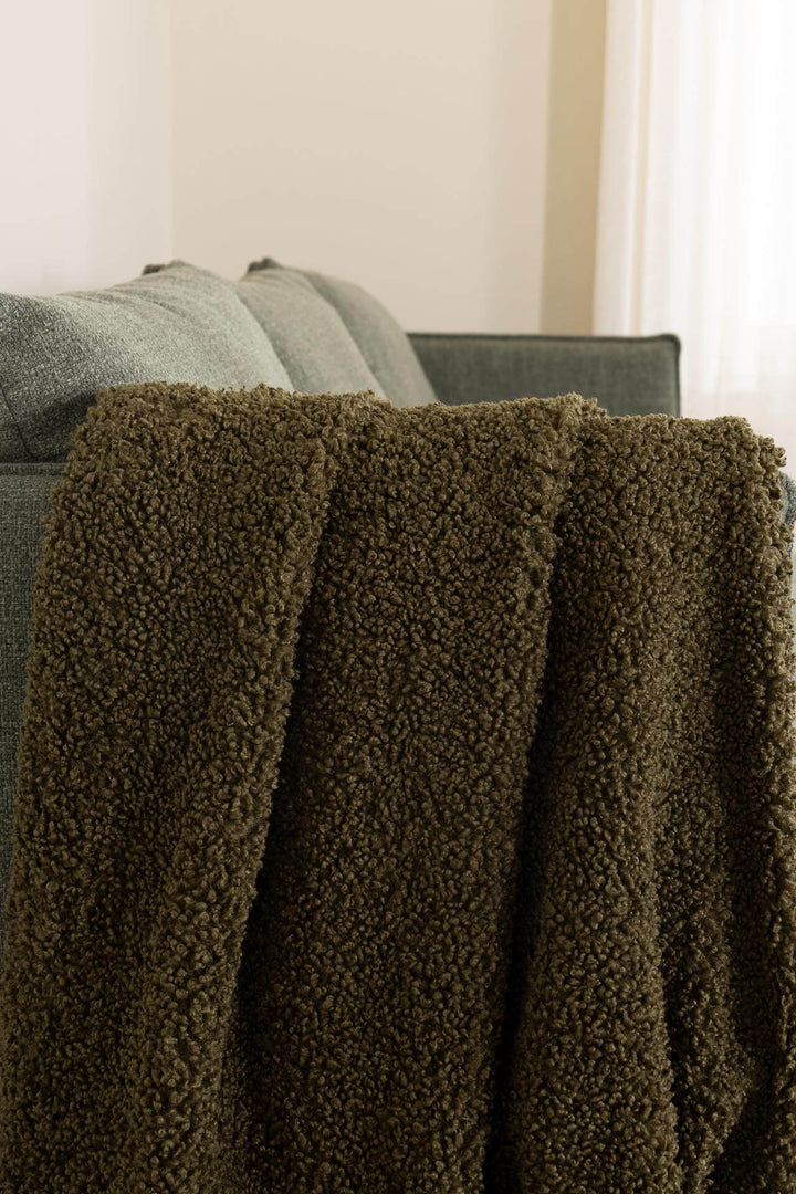 Λαδί κουβέρτα καναπέ Snug Royal Carpet με σγουρή υφή προβατάκι, ριγμένη στην πλάτη πράσινου καναπέ.
