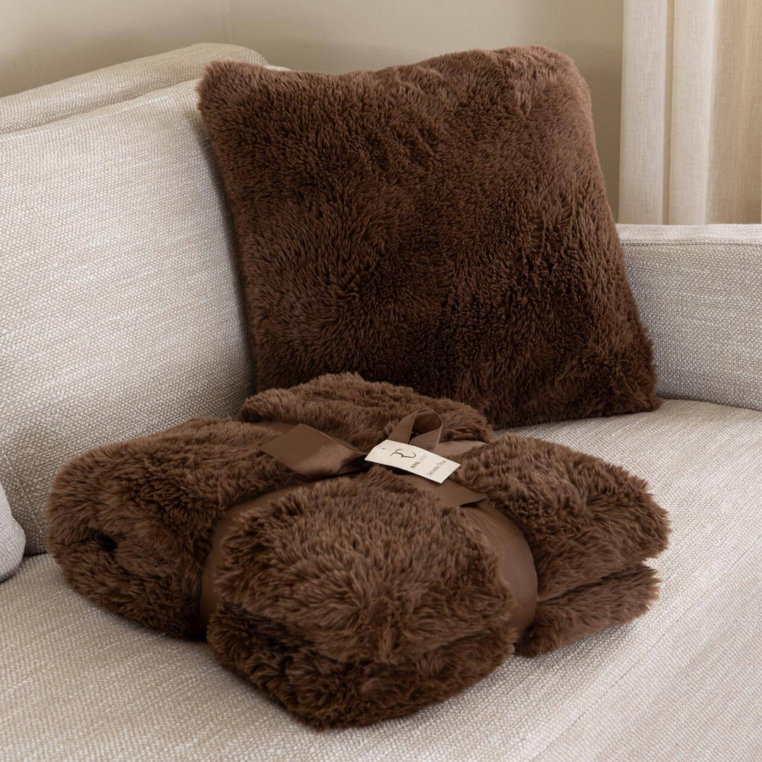 Καφέ μαξιλαροθήκη Comfy με πλούσιο γούνινο τρίχωμα πάνω σε καναπέ, δίπλα σε καφέ γούνινη κουβέρτα.