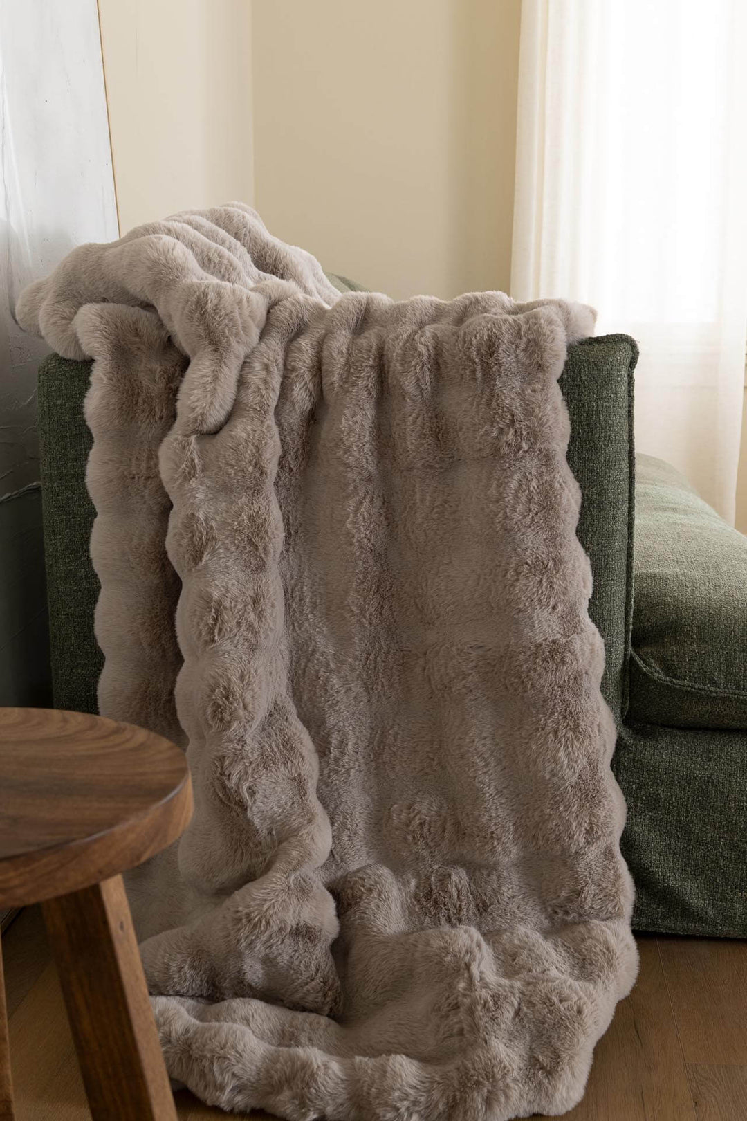 Γκρι γούνινο ριχτάρι Cosy Royal Carpet με ανάγλυφες ζώνες, ριγμένο σε πράσινη πολυθρόνα δίπλα σε ξύλινο σκαμπό.